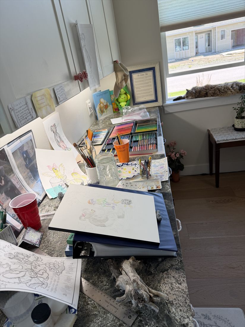 Alora's art room: a counter covered with paintings in progress, colored pencils and brushes, with Ginger the cat on the windowsill